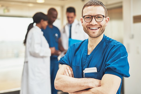 Smiling optimistic young male hospital surgeon standing in front of his colleagues with folded arms beaming at the camera
